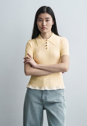 Junge Frau mit langen dunklen Haaren, die ein gelbes, kurzärmeliges, geripptes Shirt und hellblaue Jeans trägt, die Arme verschränkt, mit neutralem Gesichtsausdruck.