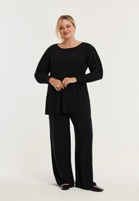Schwarzes Langarmoberteil mit rundem Halsausschnitt und gerafftem Schulterdesign, kombiniert mit locker sitzenden schwarzen Hosen und flachen schwarzen Schuhen.