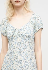 Femme portant une robe crème à imprimé floral bleu, manches ballon et un détail de nouage à l'avant sur la poitrine.