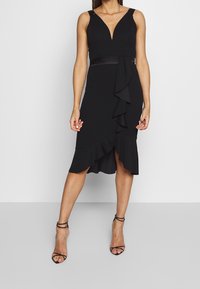 Svart klänning med V-urringning, utan ärmar och volangdetaljer. Har en figursydd siluett och en längd under knäet.