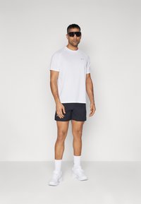 Weißes Sport-T-Shirt mit kurzen Ärmeln, kombiniert mit schwarzen Mesh-Shorts. Weiße Sportschuhe und Socken sowie Sonnenbrille runden den Look ab.