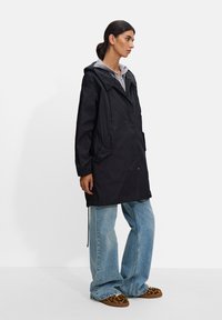 Czarna, lekka kurtka z kapturem o luźnym kroju, z dwoma bocznymi kieszeniami i regulowanymi sznurkami, noszona na szarym hoodie.