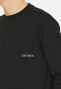 Schwarz Baumwoll Langarmshirt mit Rundhalsausschnitt und weißem Text "LES'ARCS" auf der linken Brust. Glatte Textur mit minimalen Details.