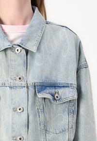 Light blue denim jacket with a relaxed fit, featuring silver snap buttons, a front pocket, and subtle "Hug" text pattern on the fabric.