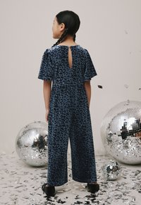 Zils leoparda raksta kombinezons ar īsām pūkainām piedurknēm un atvērumu mugurpusē, izgatavots no mīksta, teksturēta auduma, kombinēts ar melniem apaviem.