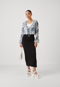 Blommig genomskinlig crop top med volangkrage, i kombination med en svartsittande midijud. Modellen bär vita platåskor och håller en vit väska.