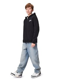 Musta vetoketjullinen huppari, jossa on etutasku ja logo. Yhdistetty vaaleansinisiin löysiin farkkuihin ja valkoisiin tennarityyppeihin. Lapsi seisoo kylki edellä.