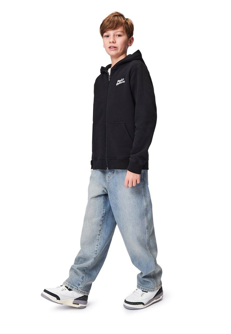 Musta vetoketjullinen huppari, jossa on etutasku ja logo. Yhdistetty vaaleansinisiin löysiin farkkuihin ja valkoisiin tennarityyppeihin. Lapsi seisoo kylki edellä.