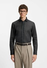 Black long-sleeve shirt with a structured collar and button closure, paired with beige pleated trousers and a brown belt. Textured fabric.