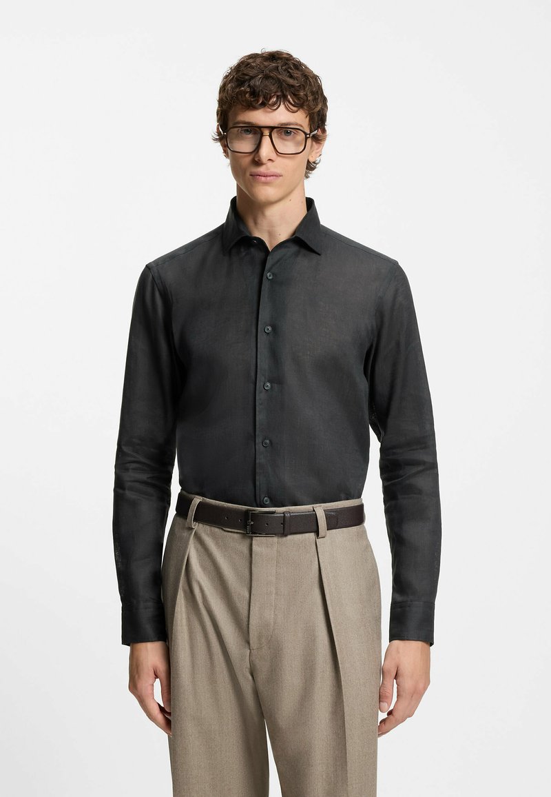 Black long-sleeve shirt with a structured collar and button closure, paired with beige pleated trousers and a brown belt. Textured fabric.