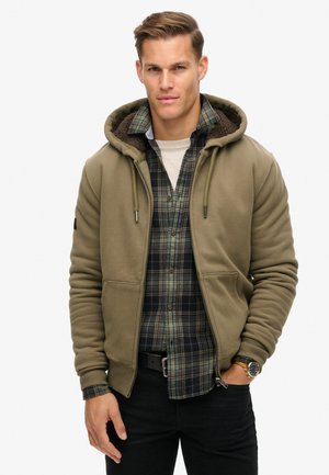 Sudadera de color verde oliva con capucha forrada de pelo, cierre de cremallera y bolsillos laterales, combinada con una camisa de cuadros. Unos vaqueros negros y un reloj completan el conjunto.