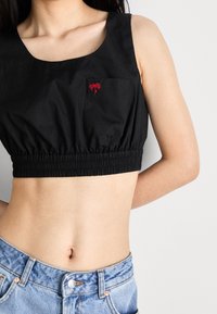 Haut court noir avec un col carré, une taille élastique et un petit logo cœur rouge sur la poche poitrine. Confectionné en tissu lisse.