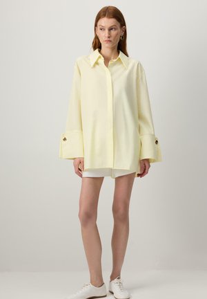 Femme aux cheveux roux portant une chemise boutonnée jaune pâle surdimensionnée et un short blanc, debout devant un fond clair uni.
