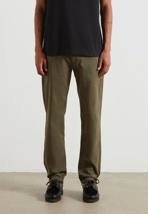 Osoba ubrana w oliwkowozielone spodnie o prostych nogawkach, czarne sznurowane buty oraz czarną koszulę, stojąca na jasnej podłodze na tle białej ściany.