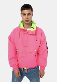 Rosa übergroße Jacke mit einer neon-grünen Kapuze, verstellbaren Kordeln, Druckknöpfen und einer vorderen Klapptasche. Text auf dem Ärmel.