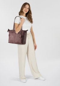 Bolso tote de cuero burdeos con superficie texturada, dos asas superiores y una etiqueta extraíble. Modelo lleva una camisa blanca y pantalones anchos color crema.
