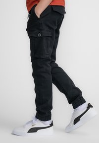 Zwarte cargobroek met zijzakken, taps toelopende manchetten en een gladde textuur. Gepaard met witte sneakers met zwarte accenten en logo.