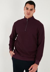 Suéter burdeos de cuello con cremallera y textura acanalada, con mangas largas y puños ajustados. Cuenta con una cremallera corta en el cuello para ponérselo con facilidad.