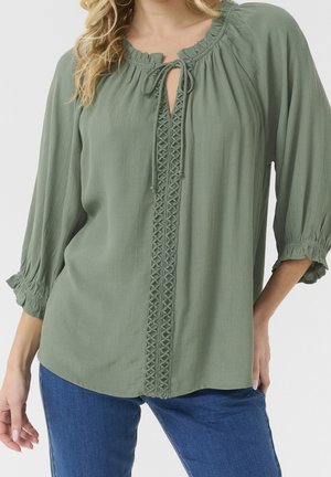 Žena oblečená v šalvějově zelené blůze se shrnovaným výstřihem, předním zavazováním, dekorativním svislým lemováním a tříčtvrtečními rukávy, kombinovaná s modrými džíny.