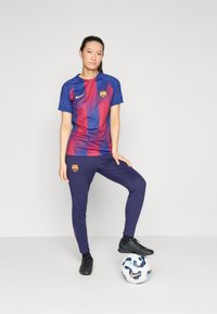 Barcelona FC-tröja med blå och röda vertikala ränder, kombinerad med marinblå byxor som har lagets logotyp; bärs med svarta fotbollsskor och en fotboll.