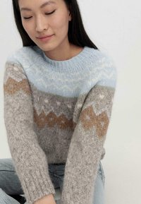 Neulepusero, jossa on sinisiä, harmaita ja ruskeita geometrisia kuvioita. Pehmeä, teksturoitu kangas, jossa on pyöreä pääntie ja pitkät hihat.