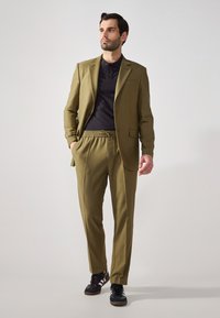 Completo verde oliva con un design snello; blazer a un bottone e pantaloni dal taglio morbido con cintura elasticizzata; abbinato a sneaker nere.