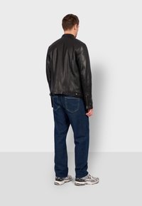 Chaqueta de cuero negro con un ajuste slim, que presenta una textura suave y cremalleras discretas, combinada con pantalones vaqueros azules y deportivas.