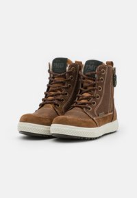 Botas de tobillo de ante marrón con ojales de metal, cordones marrones, una suela de goma blanca texturizada y un logo de parche en la lengüeta.
