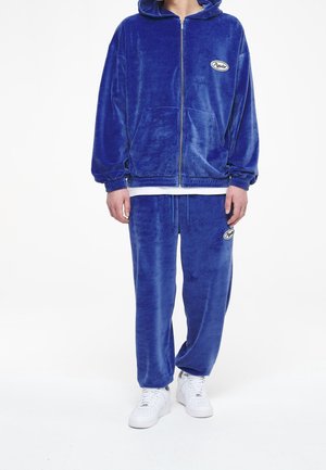 Set di felpa con cappuccio e pantaloni tuta in blu brillante, caratterizzato da una patch con logo ricamato. Il materiale ha una consistenza morbida, con polsini elasticizzati.
