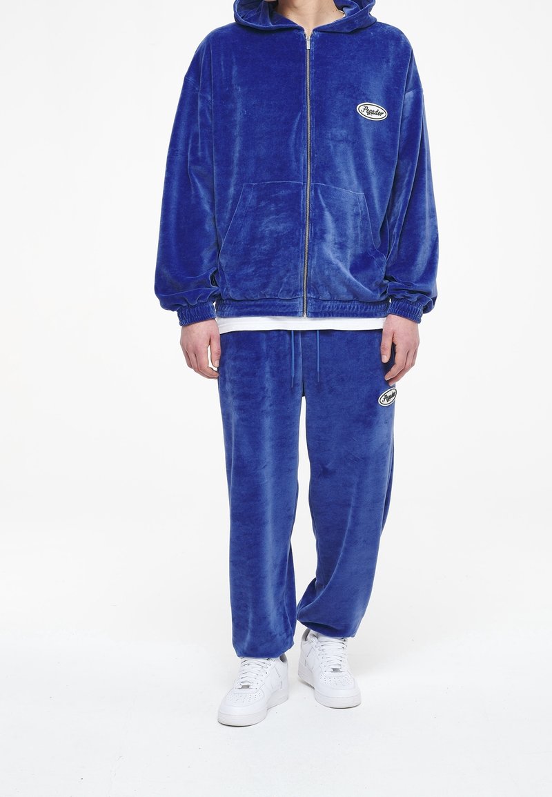 Conjunto de sudadera con capucha y pantalones de chándal de terciopelo en un azul brillante, con un parche de logo bordado. El material tiene una textura suave y cuenta con puños elásticos.