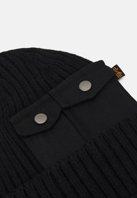 Black knit beanie with ribbed texture, featuring a flat, canvas band with snap buttons and a small yellow tag detail.