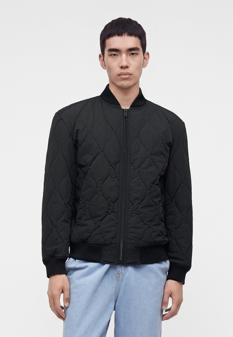 Jeune homme aux cheveux courts noirs portant une veste bomber matelassée noire et un jean bleu clair, debout devant un fond gris uni.