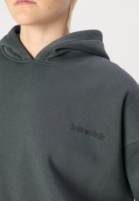 Sudadera de color verde oscuro con una textura gruesa. Presenta un bolsillo frontal y capucha, mostrando "la vie est belle" bordado en un hilo de contraste.