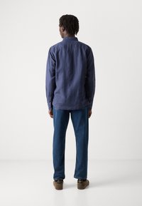 Marineblå skjorte med lange ermer laget av teksturert materiale, kombinert med løse denimbukser og brune sko, vist bakfra.
