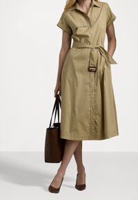 Khaki shirt dress with rolled short sleeves, button front, tie waist, and A-line skirt. Brown tote bag and matching heels complete the look.