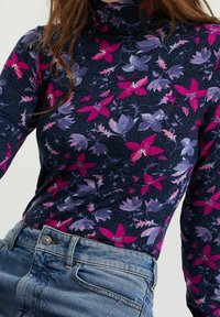Kvinna iklädd en figurnära marinblå polotröja med rosa och lila blommönster, nedstoppad i ljusblå denimjeans.