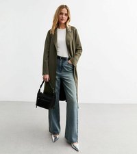 Trench coat verde oliva, top bianco, jeans in denim a vita alta, scarpe argentate a punta, borsa nera, in piedi su un pavimento grigio chiaro.