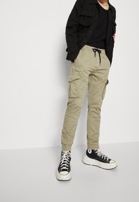 Khaki cargo-byxor med elastiska muddar, med sidofickor och dragsko i midjan, som kombineras med svarta högtoppsneakers.