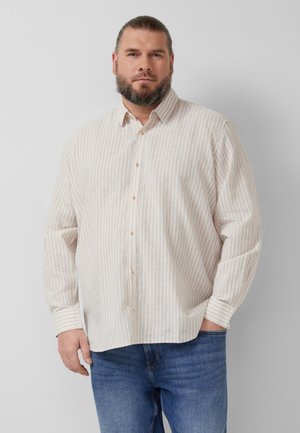 Mann mit Bart, der ein beige gestreiftes Hemd mit Knöpfen und blaue Jeans trägt, steht vor einer schlichten hellen Wand.