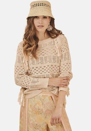 Sweater de punto en beige claro con patrón calado y acentos de flecos; combinado con pantalones de patrón floral y un sombrero de cubo de tejido.