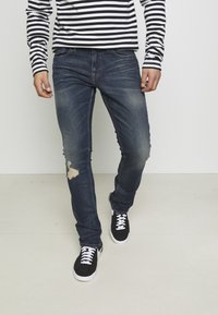 Modré džíny z denimu s úzkým střihem a natrženým detailem na koleni. Kombinované s černými teniskami a černobílým pruhovaným dlouhým rukávem.