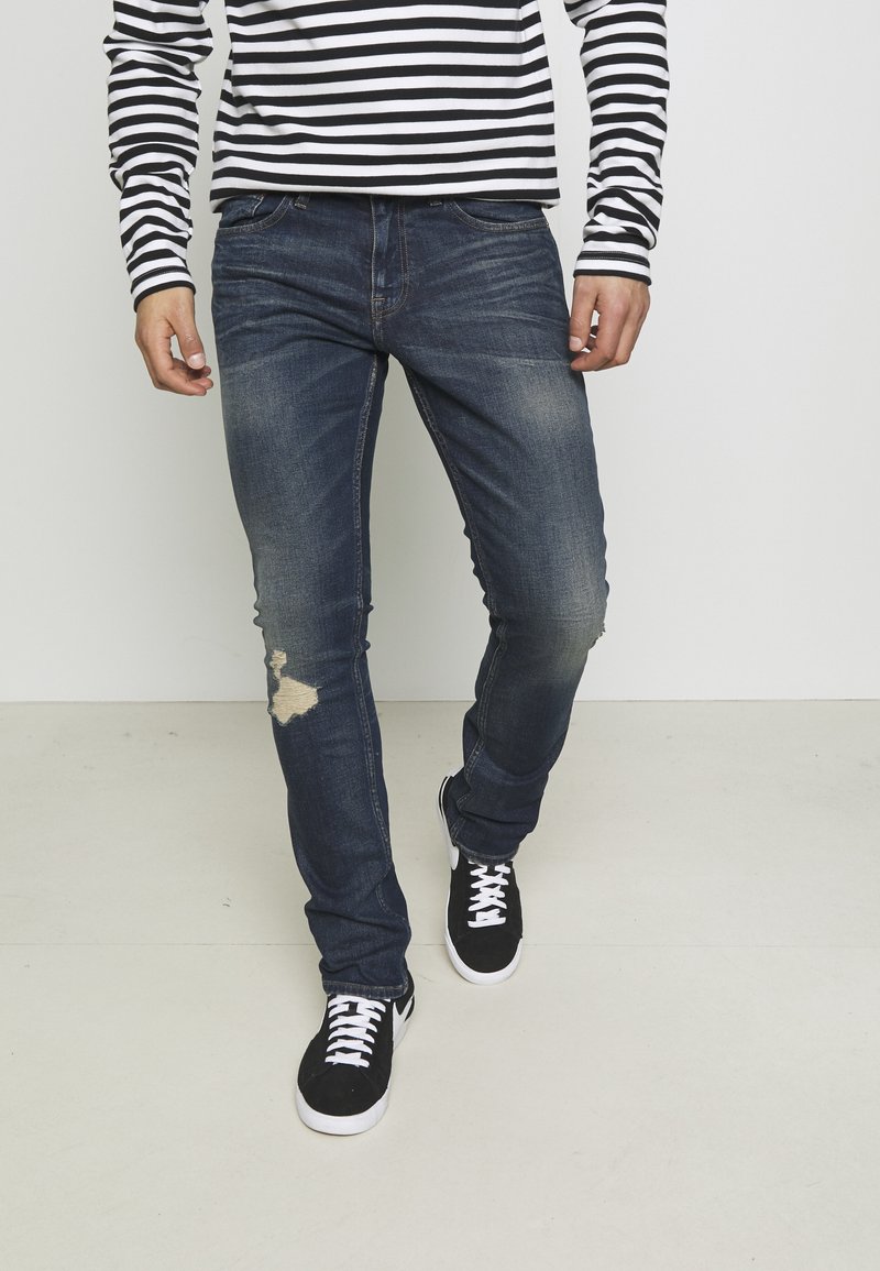 Modré džíny z denimu s úzkým střihem a natrženým detailem na koleni. Kombinované s černými teniskami a černobílým pruhovaným dlouhým rukávem.