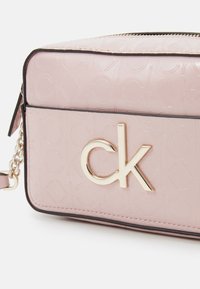 Jasnoróżowa prostokątna torebka na ramię z wytłaczanym wzorem "ck" i dużym złotym logo "ck" na przedniej kieszeni, z detalem złotego łańcuszka na pasku.