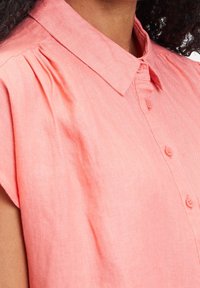 Nainen, joka pukeutuu korallinpunaisiin lyhythihaiseen puseroon, jossa on napit ja kaulus. Puseron pinta on kuvioitu ja olkapään lähellä on hienoja laskoksia.