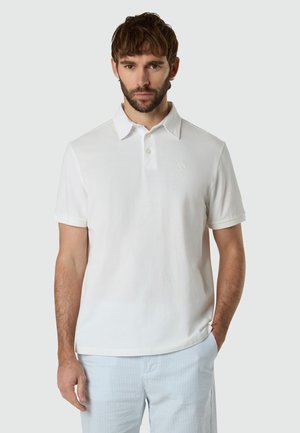Hombre de cabello castaño y barba que lleva una camisa polo blanca de manga corta y pantalones azul claro con rayas, de pie frente a un fondo gris claro liso.