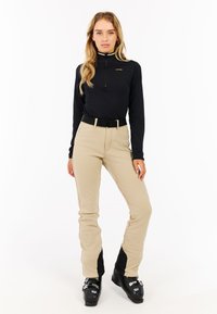 Sort zip-up trøje med guld detaljer, parret med lysebrune bukser med en sort accent forneden, båret med sorte skistøvler og et sort bælte.