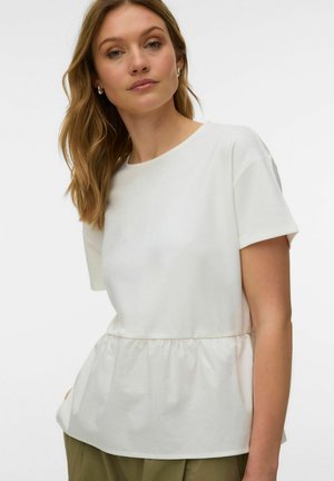 Femme aux cheveux châtain clair portant des boucles d'oreilles argentées en forme de créole et un haut blanc à manches courtes style péplum, avec un pantalon vert olive, sur fond blanc.