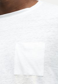 Κοντινό πλάνο ενός ατόμου που φοράει λευκό T-shirt από υφασμάτινη ύλη με μικρή τσέπη στο στήθος και στρογγυλή λαιμόκοψη.