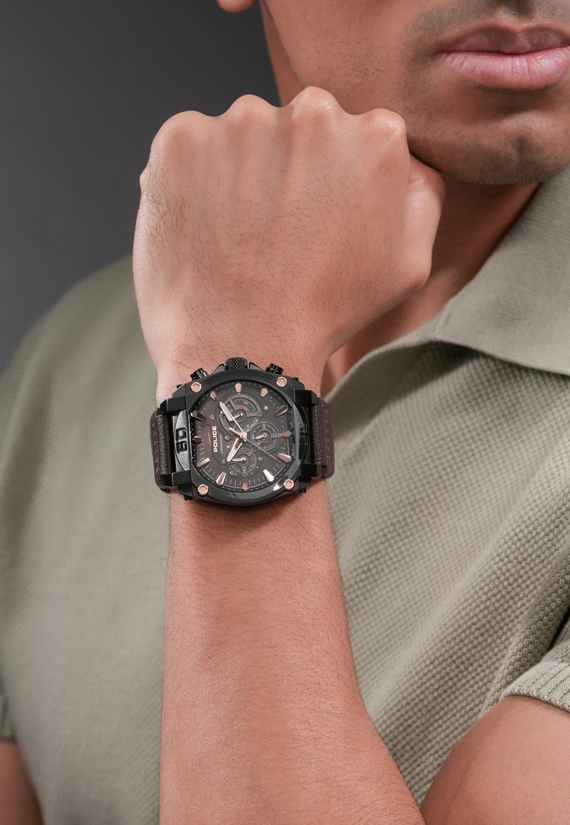 Hombre con un reloj cronógrafo de pulsera negro con correa de cuero marrón, apoyando su puño cerrado contra el mentón.