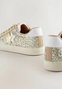 Mirdzoši zelta un sudraba sporta apavi ar baltu un smilškrāsas dizainu. Iekļauj zvaigžņu akcentus, teksturētu augšdaļu un gumijas zoli.